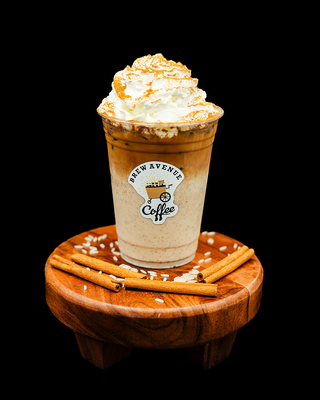 A clear cup with a sticker showing the Brew Avenue Coffee logo is on a wooden stool against a black studio background. The cup is filled with Cafe Horchata with white foam on top and syrup drizzled on top of the foam. Cinnamon sticks and white rice are sprinkled on the stool.