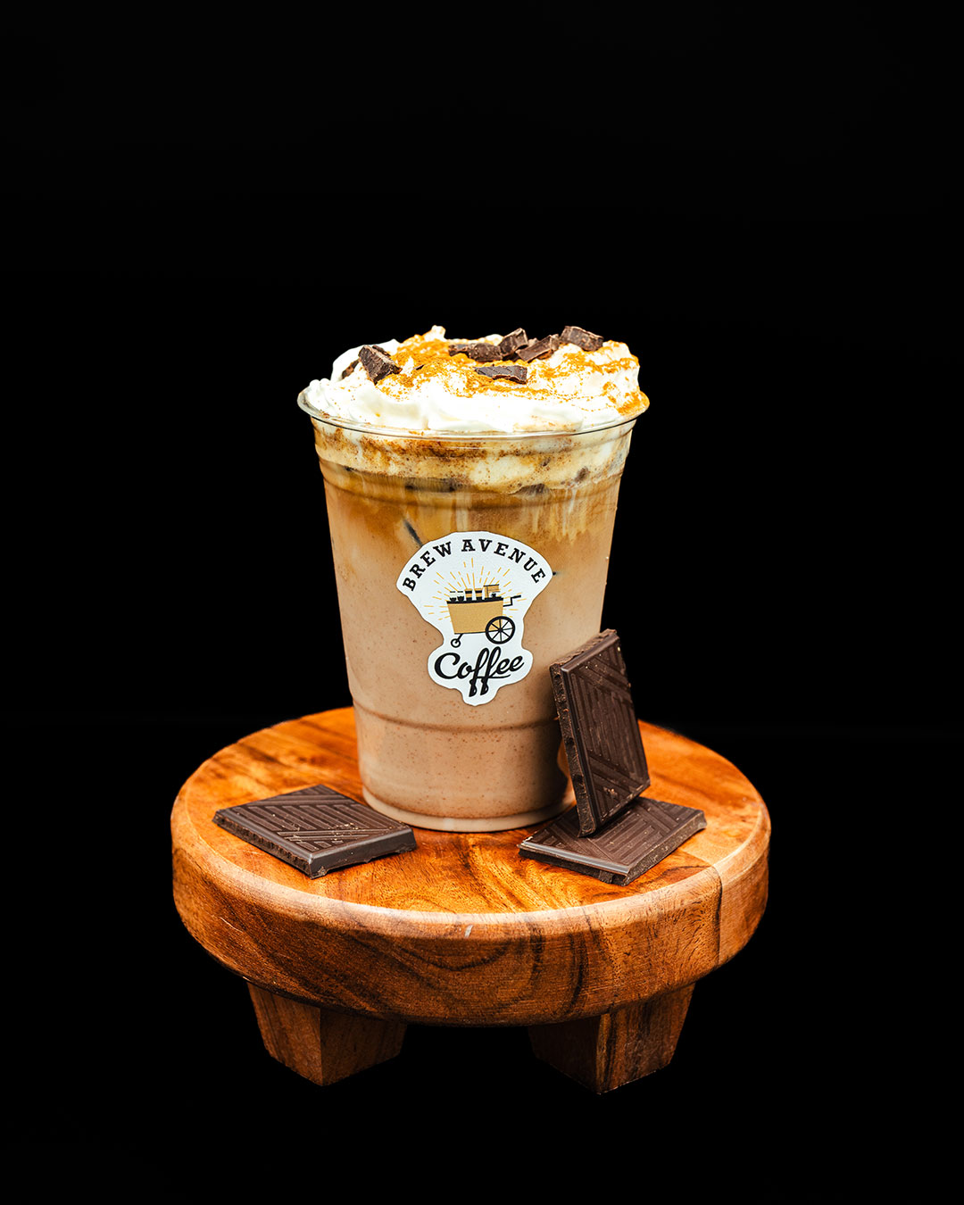 A clear cup with a sticker showing the Brew Avenue Coffee logo is on a wooden stool against a black studio background. The cup is filled with cafe mocha with cream and chocolate shavings on top. Chocolate squares are also on the stool.