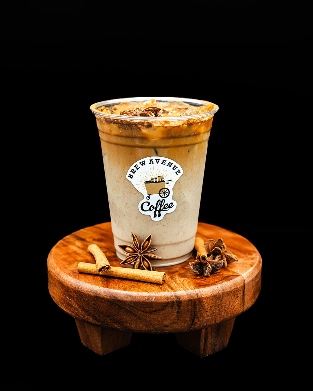A clear cup with a sticker that shows the Brew Avenue Coffee logo is sitting on a wooden stool against a black studio background. The cup is filled with iced chai latte with cinnamon sticks on top of the stool.