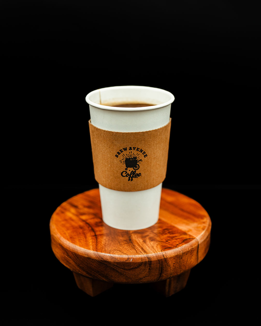 A white cup with a sleeve that shows the Brew Avenue Coffee logo is sitting on a wooden stool against a black studio background. The cup is filled with a hot Americano.