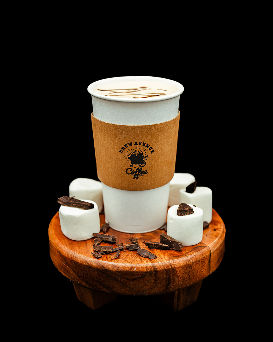 Hot Chocolate in a white cup with the Brew Avenue Coffee logo on a wooden stool. Marshmallows with chocolate chips and shavings also sit on top of the stool.