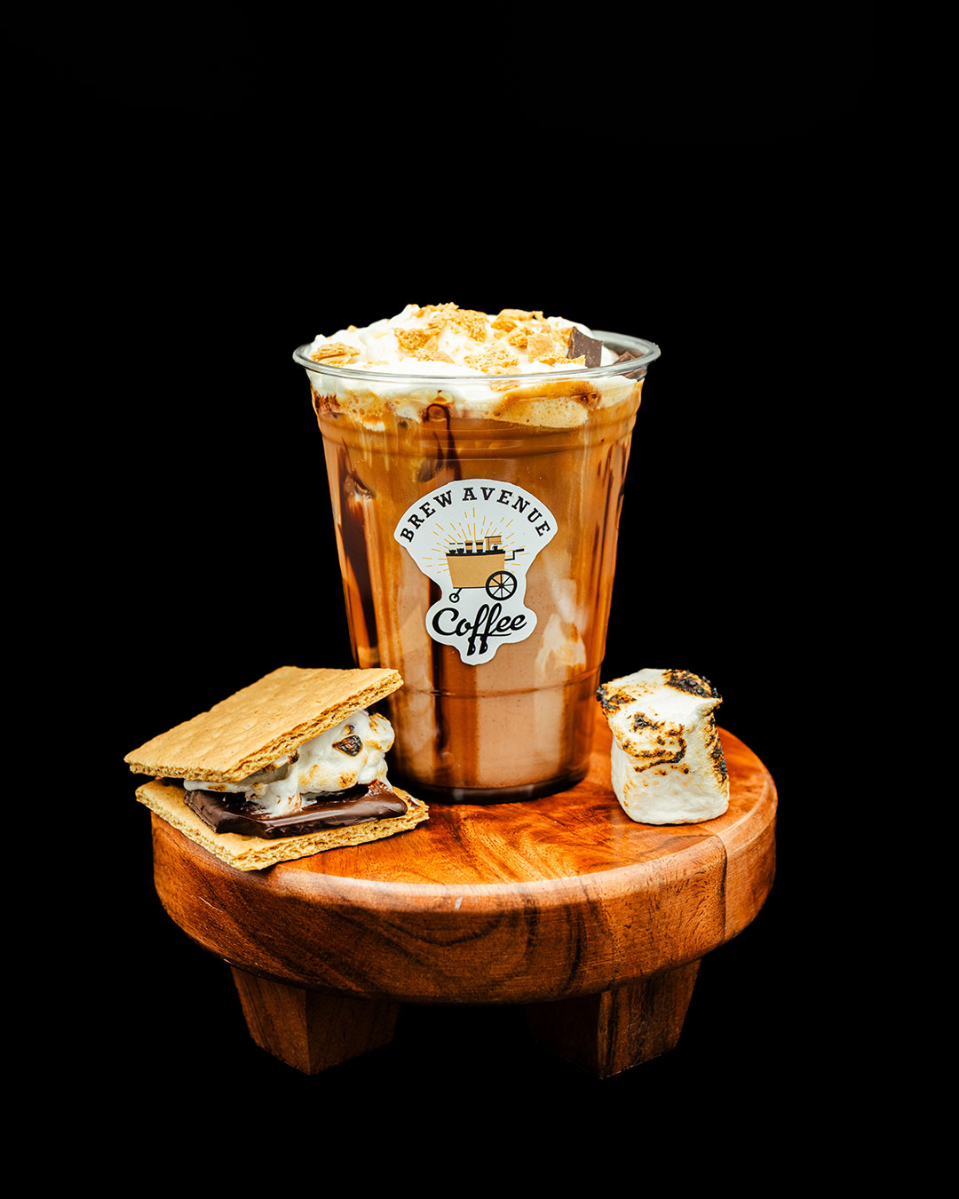 A clear cup with a sticker showing the Brew Avenue Coffee logo is on a wooden stool against a black studio background. The cup is filled with toasted marshmallow coffee drink with syrup drizzling down the sides. Smore and toasted marshmallow is also on the stool.