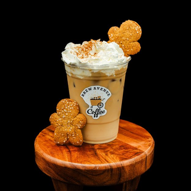 A clear cup with a sticker showing the Brew Avenue Coffee logo is on a wooden stool against a black studio background. The cup is filled with Gingerbread latte with white foam on top and gingerbread man on top of the foam. Gingerbread man also on the stool.