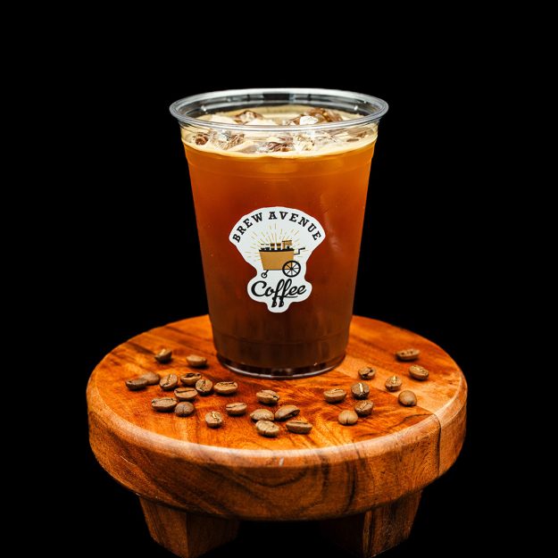A clear cup with a sticker that shows the Brew Avenue Coffee logo is sitting on a wooden stool against a black studio background. The cup is filled with iced americano with cofee beans on top of the stool.