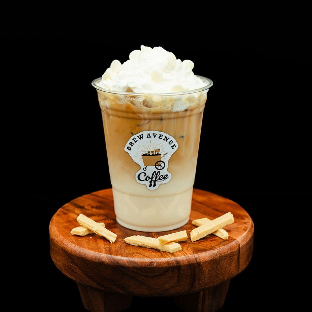 A clear cup with a sticker that shows the Brew Avenue Coffee logo is sitting on a wooden stool against a black studio background. The cup is filled with Macadamia Nut Sweet Cream coffee with white foam on top.