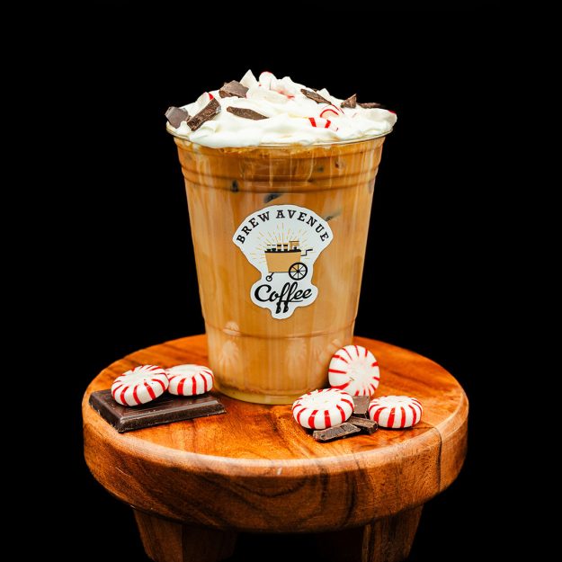A clear cup with a sticker showing the Brew Avenue Coffee logo is on a wooden stool against a black studio background. The cup is filled with peppermint mocha with cream peppermints and chocolate on top. Peppermints and chocolate are also on the stool.