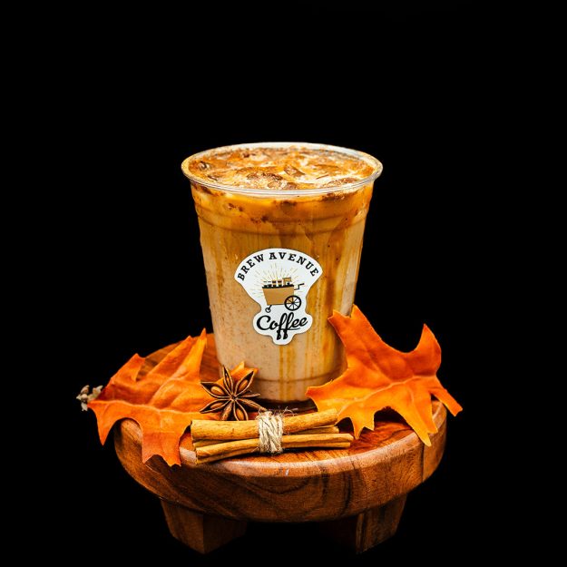 A clear cup with a sticker showing the Brew Avenue Coffee logo is on a wooden stool against a black studio background. The cup is filled with pumpkin chai with ice. Fall leaf and cinnamon sticks are also on the stool.
