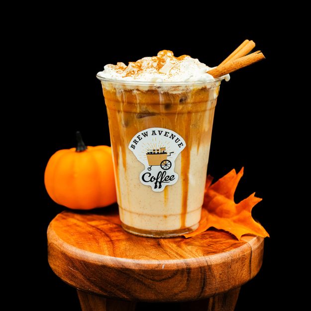 A clear cup with a sticker showing the Brew Avenue Coffee logo is on a wooden stool against a black studio background. The cup is filled with pumpkin pie latte with white foam on top and cinnamon sticks with cinnnamon dust on top of the foam. Fall leaf and pumpkin are also on the stool.