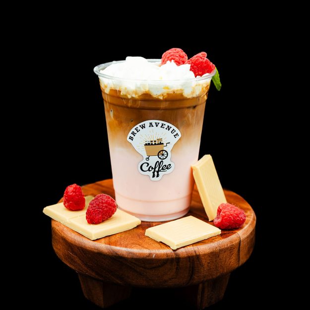 A clear cup with a sticker showing the Brew Avenue Coffee logo is on a wooden stool against a black studio background. The cup is filled with Raspberry white mocha coffee drink with white foam on top and raspberries with a green mint on top of the foam. White chocolate and raspberries are also on the stool.