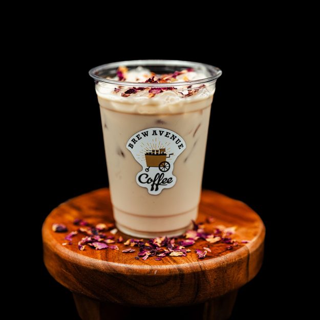 A clear cup with a sticker that shows the Brew Avenue Coffee logo is sitting on a wooden stool against a black studio background. The cup is filled with Rose Chai with rose petals on top of the drink and stool.