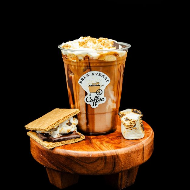 A clear cup with a sticker showing the Brew Avenue Coffee logo is on a wooden stool against a black studio background. The cup is filled with toasted marshmallow coffee drink with syrup drizzling down the sides. Smore and toasted marshmallow is also on the stool.