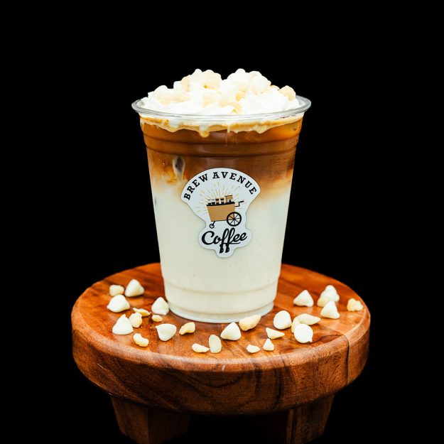 A clear cup with a sticker showing the Brew Avenue Coffee logo is on a wooden stool against a black studio background. The cup is filled with white mocha coffee drink that's white on the bottom, brown coffee on top with white foam. white chocolate drops are also on the stool.